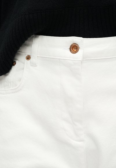 Κοντινό πλάνο λευκών τζιν από τζιν ύφασμα με χάλκινο κουμπί και νίκελ, σε συνδυασμό με μαύρο ριπ πουλόβερ που έχει φορεθεί μέσα.