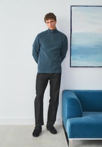 Maglione zip-up blu scuro in tessuto testurizzato, pantaloni neri e scarpe nere. Sullo sfondo si vede un divano in velluto blu e un quadro astratto.