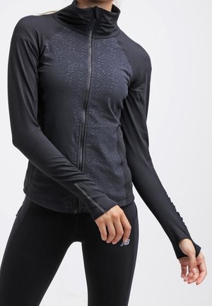 Frau trägt eine schwarze, langärmelige Sportjacke mit Reißverschluss und schwarze Leggings mit kleinem Logo am Oberschenkel vor weißem Hintergrund.