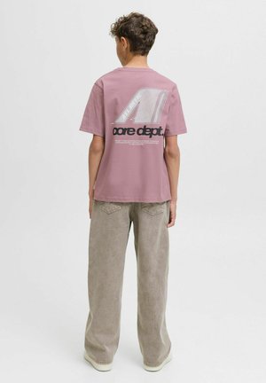 Una persona con capelli ricci di spalle, indossa una maglietta rosa con un logo grafico sulla schiena e pantaloni larghi e leggeri.