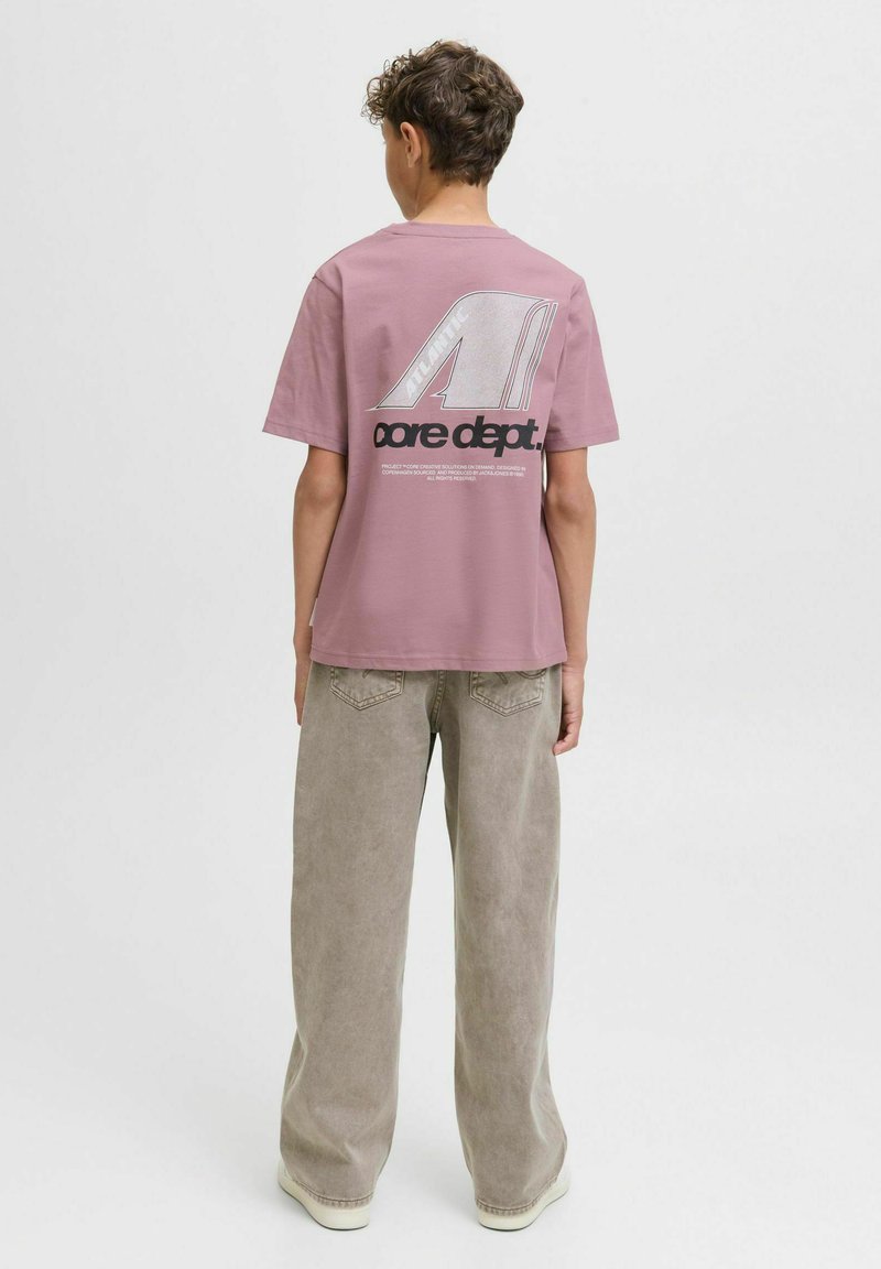 Een persoon met krullend haar staat met zijn rug naar ons toegekeerd, draagt een roze shirt met een grafische logo op de achterkant en lichte, wijde broek.