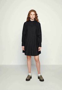 Vestido preto com botões à frente, laterais franzidas e mangas longas. Usado com sapatos verde-oliva de slip-on e meias brancas.