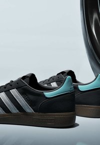 Baskets Adidas noires avec des bandes blanches et des accents bleu sarcelle au talon sur une surface grise, partiellement en miroir sur le côté droit.