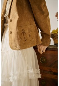 Veste en daim marron avec broderie florale sur la poche avant portée sur une jupe en tulle blanche, main reposant sur une commode en bois avec un panier de fruits.