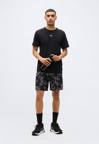 Νεαρός άνδρας με μαύρο μπλουζάκι Puma, σορτς παραλλαγής, μαύρα αθλητικά παπούτσια και κάλτσες, κρατώντας μαύρο παγούρι, μπροστά σε απλό φόντο.