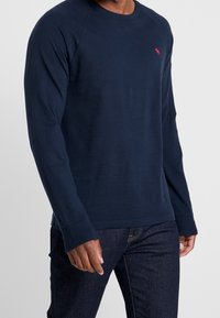 Marineblaue Longsleeve Crewneck Sweatshirt aus Baumwolle, mit gerippten Bündchen und einem kleinen gestickten Logo auf der Brust.