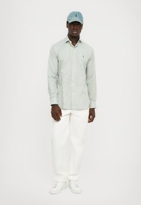 Cămașă button-up cu mâneci lungi de culoare verde deschis, cu un logo mic, asortată cu pantaloni albi și adidași albi. Pălăria este din denim deschis, cu logo.