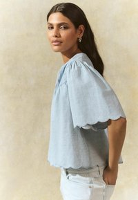 Light blue scalloped hem blouse made of soft fabric, featuring puff sleeves and a round neckline, styled with light denim jeans.
