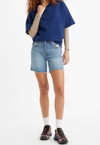 Modrý oversized svetr, béžová pruhovaná košile s dlouhým rukávem pod ním, světle modré denimové šortky s třepenými okraji, doplněné hnědými sportovními botami.