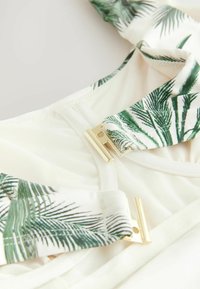 Sangles ajustables à motif de palmiers en vert et blanc, avec des accessoires en métal doré, sur une base en tissu de couleur claire avec une finition texturée.