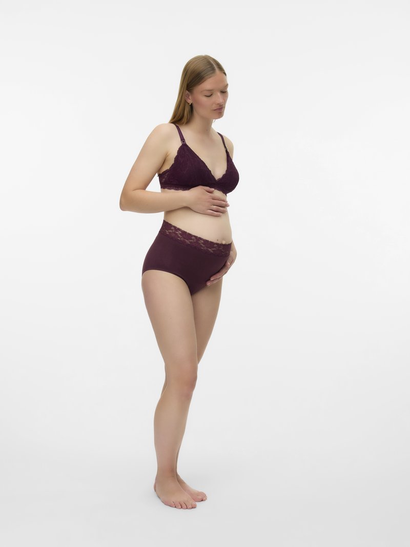 Έγκυος γυναίκα με μπορντό δαντελένιο σουτιέν και αντίστοιχο ψηλόμεσο εσώρουχο, που κρατά απαλά την κοιλιά της, στέκεται ξυπόλυτη σε λευκό φόντο.