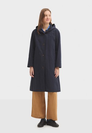 Manteau bleu marine jusqu'aux genoux avec capuche, fermeture à boutons à l'avant et manches longues. Porté avec un pantalon large couleur beige et des chaussures noires.