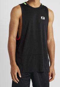 Lihaksikas henkilö, jolla on päällä musta hihaton Nike-toppi, jossa on leveät kädenaukot, sekä mustat urheilushortsit yksinkertaista taustaa vasten.