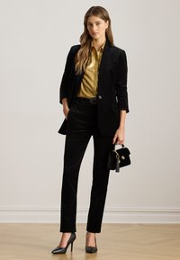 Blazer e pantaloni in velluto nero abbinati a una blusa in satin oro. Gli accessori includono una piccola borsa nera e tacchi grigi.