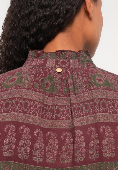 Burgundijas blūze ar ziedu rakstu, ar viļņotu apkakli un pogu detaļu aizmugurē. Izveidota no teksturētas auduma ar zaļiem un baltiem akcentiem.