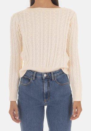 Kvinde iført cremefarvet kabelstrikket sweater med bådudskæring og blå højtaljede jeans, stående med armene afslappet ned langs siderne.