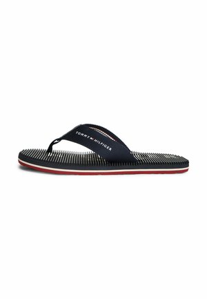Une tong rayée noire et grise avec une semelle rouge et une bride bleu marine, marquée "Tommy Hilfiger" en texte blanc.