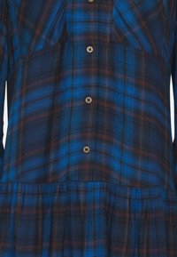 Blå rutig skjorta med knäppning, med brun och svart accent. Tillverkad av texturerad tyg med en samlad nederdel.