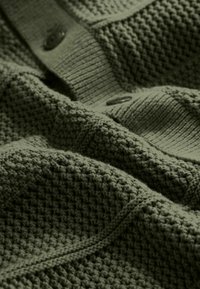 Gros plan sur un tissu tricoté vert olive texturé avec un détail côtelé et des boutons visibles.
