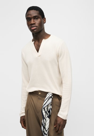 Giovane uomo che indossa una maglia a maniche lunghe color crema con scollo a V e pantaloni marroni, con una sciarpa a motivo bianco e nero infilata nella cintura.