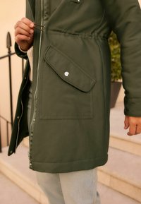 Olivgrüne Parkajacke mit Reißverschluss, taillierter Taille und Fronttasche mit Druckknopfverschluss. Glattes Material und minimale Designdetails.