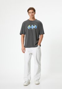 Camiseta de algodón gris con un logo colorido de Batman en la parte delantera, combinada con pantalones de mezclilla blancos y zapatillas blancas. Ajuste simple y relajado.