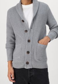 Grå stickad cardigan med sjalkrage, fem bruna knappar och två framfickor. Lättviktsmaterial och ribbade manschetter.