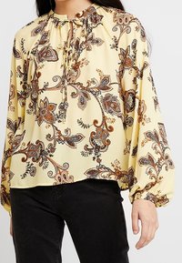Long-sleeve blouse in pastel yellow fabric with a floral paisley pattern in brown and orange. Features a gathered neckline and tie detail.