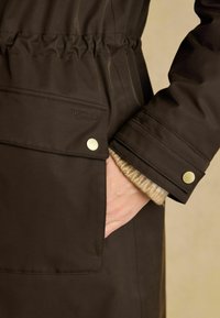  Käsi on paigutatud tumeda pruuni jope taskusse, millel on kuldsed nööbid ja nähtav beež kudum cuffs all. 