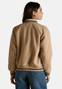 Kreemjas fleece bomber jakk, millel on ribatud kaelus ja mansetid, ning millel on triibuline aksentlint. Tagumiselt vaates on näha mugav lõige ja tekstuurne kangas.