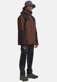 Wasserabweisende braune und schwarze Jacke, grauer Sonnenhat, schwarze Hose und graue Wanderschuhe; mit Reißverschlüssen und verstellbaren Manschetten.