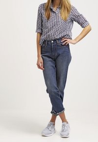 Blå denimjeans med avslappnad passform och uppvikt nederkant, tillsammans med en mönstrad svart och vit kortärmad skjorta och grå sneakers.