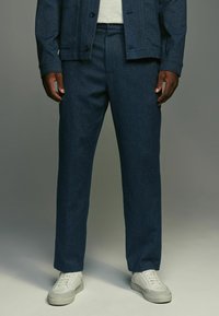 Pantaloni blu scuro dal design su misura con una texture liscia, abbinati a sneakers bianche, caratterizzati da gamba dritta e pieghe sottili.