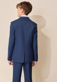 Niño con cabello castaño claro vistiendo un traje azul a cuadros y camisa blanca, de pie de espaldas sobre un fondo beige.