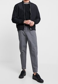 Giubbotto bomber nero con zip, abbinato a una maglietta nera e pantaloni grigi a gamba affusolata. Sneakers nere completano l'outfit.