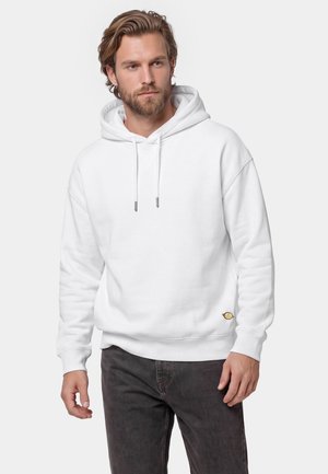 Mand med lyst brunt hår og skæg iført en ensfarvet hvid hættetrøje og mørke jeans, stående mod en lys baggrund.