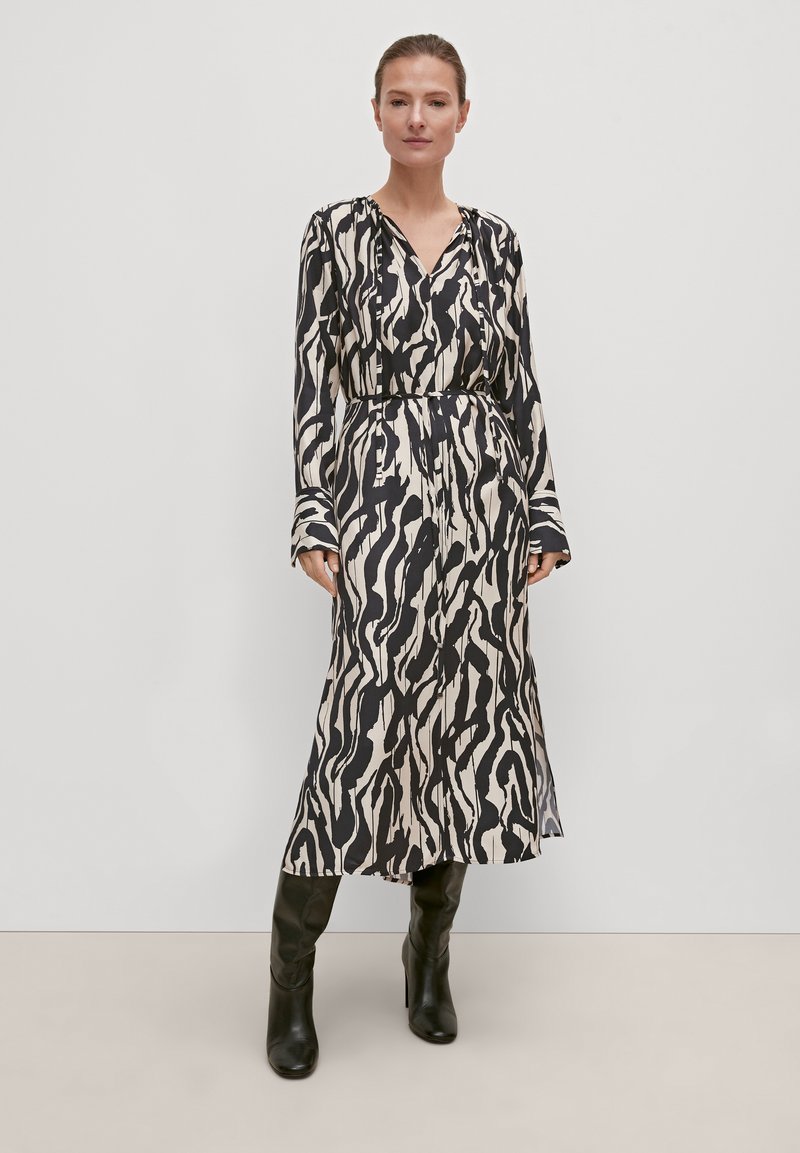 Langarmkleid mit einem schwarzen und cremefarbenen abstrakten Muster, V-Ausschnitt, tailliertem Bund und fließender Silhouette, kombiniert mit knielangen schwarzen Stiefeln.