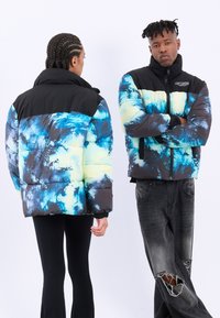Puffjakke med sort top, blå og gul tie-dye mønster på den nederste halvdel, lynlåslukning, elastiske manchetter, båret med sorte jeans og leggings.