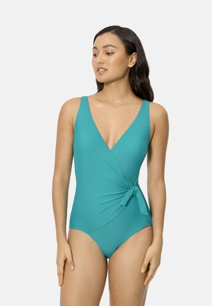 Mujer con traje de baño de una pieza color verde azulado sin mangas, con escote en V y lazo lateral, de pie contra un fondo claro y simple, mirando hacia un lado.