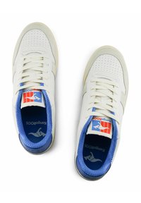 Baskets blanches avec un bout perforé, doublure intérieure bleue et lacets. Présentant un écusson bleu et rouge sur la languette et une semelle en caoutchouc texturée.