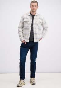 Geruit flanellen overhemd in wit, beige en grijs, voorzien van twee borstzakken en knoopsluiting, gecombineerd met donker denim en beige sneakers.