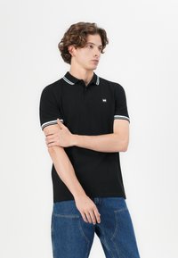 Black polo shirt with white striped collar and sleeves, featuring a small sewn logo on the chest; smooth fabric and classic fit.