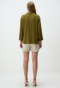 Blusa verde oliva a maniche lunghe con colletto, polsini abbottonati e una texture liscia; abbinata a pantaloni corti beige chiaro e sandali con tacco.