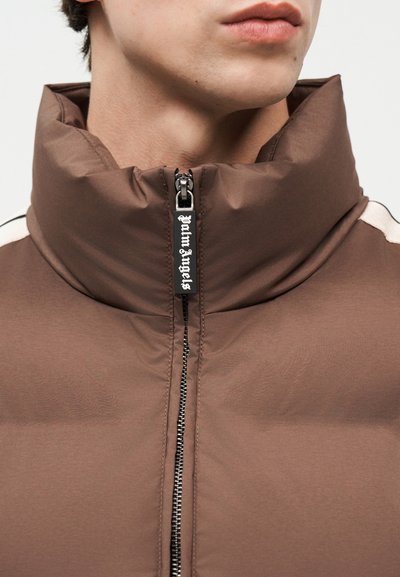 Nahaufnahme einer Person, die eine braune gesteppte Jacke mit hohem Kragen trägt, auf dem ein schwarzes Sichtlabel am Reißverschluss mit der Aufschrift "Palm Angels" zu sehen ist.