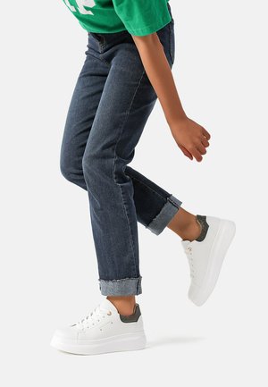 Sneaker bianche con un accento grigio testurizzato sul tallone, suola piatta e punta rotonda, abbinate a jeans scuri in denim risvoltati.