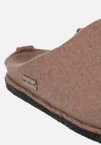 Bruine vilten pantoffel met een zwarte rubberen zool, met een flatte, ronde vorm en een klein metalen merklabel aan de zijkant.