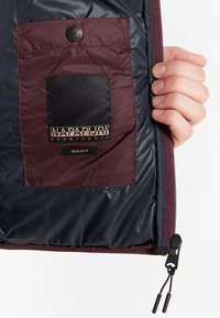 La giacca isolata maroon e navy presenta all'interno una tasca nera con etichetta logo e una chiusura a zip con un tirante in tessuto.