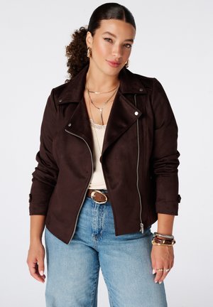 Vrouw met krullend haar in een paardenstaart, die een donkerbruine jas draagt over een wit topje, blauwe spijkerbroek, gouden sieraden en een bruine riem.