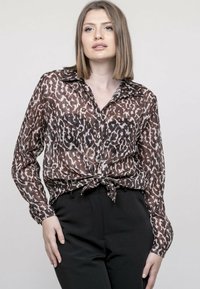 Blouse à imprimé léopard dans des tons de marron et de beige, avec des manches longues, un col, une fermeture à boutons sur le devant, et un détail de taille nouée.