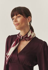 Eșarfă din mătase în nuanțe de burgund și crem, cu un model geometric, legată la gât, asortată cu cercei triunghiulari aurii.